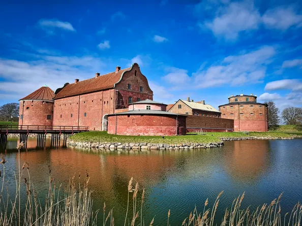 En historisk byggnad med röd tegelvägg och rundade torn, belägen vid en spegelblanka sjö under en klar blå himmel.
